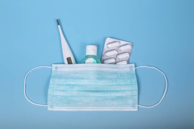 Tedavi için koruma ürünleri. Cerrahi maske, termometre, dezenfektan jeli ve mavi arka planda hastalık önleyici ilaçlar. Coronavirus veya covid-19 konsepti.