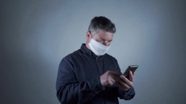 Gri arka planda akıllı telefon kullanarak internette gezinen koruyucu maskeli çekici genç bir adam.