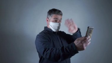 Çekici adam gri arka planda akıllı telefonla görüntülü sohbet için koruyucu maskeyi çıkarıyor.
