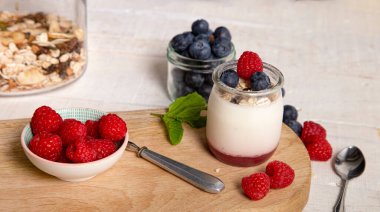 Kapalı cam kavanoz dolusu beyaz yoğurt, taze ahududu ve yabanmersini servis masasında, arka planda ahşap tepsi, kopyalama alanı..