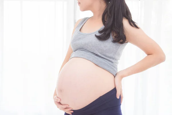 Abgeschnittenes Bild einer schwangeren Frau, die ihren Bauch vor dem Fenster hält — Stockfoto