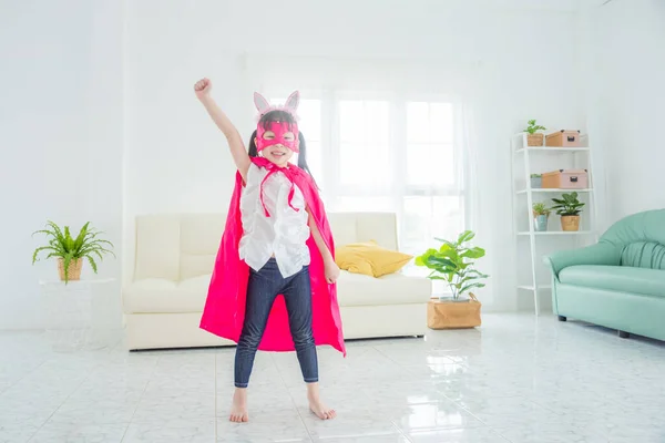 Gelukkig klein aziatisch meisje dragen roze superheld kleding staan op woonkamer vloer Stockfoto