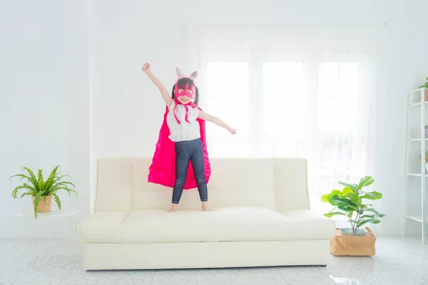 Gelukkig klein aziatisch meisje dragen roze superheld kleding staan op woonkamer bank Stockfoto
