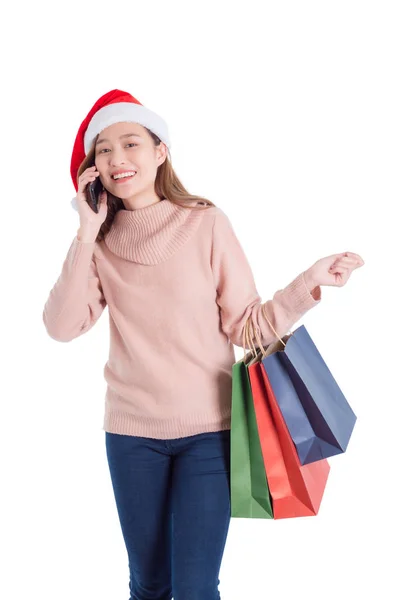 Mooi aziatisch meisje dragen santa claus hoed, het dragen van vele papieren boodschappentassen en praten via smartphone — Stockfoto