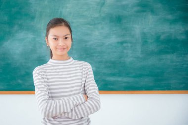 Asyalı genç bir kız öğrenci ayakta duruyor ve yeşil karatahtanın önünde gülümsüyor..