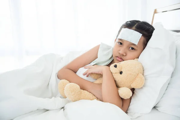 Asian sick girl covered in blanket is hugging teddy bear with sad face while lying on bed.