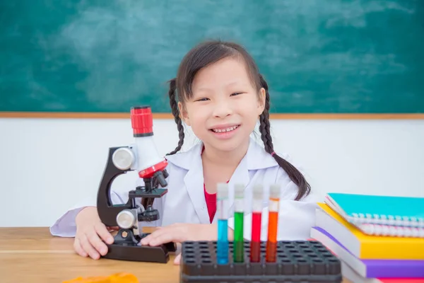 白いコートを着たアジア系の学生が 理科教室の黒板の前でドレックと笑顔で座っています — ストック写真
