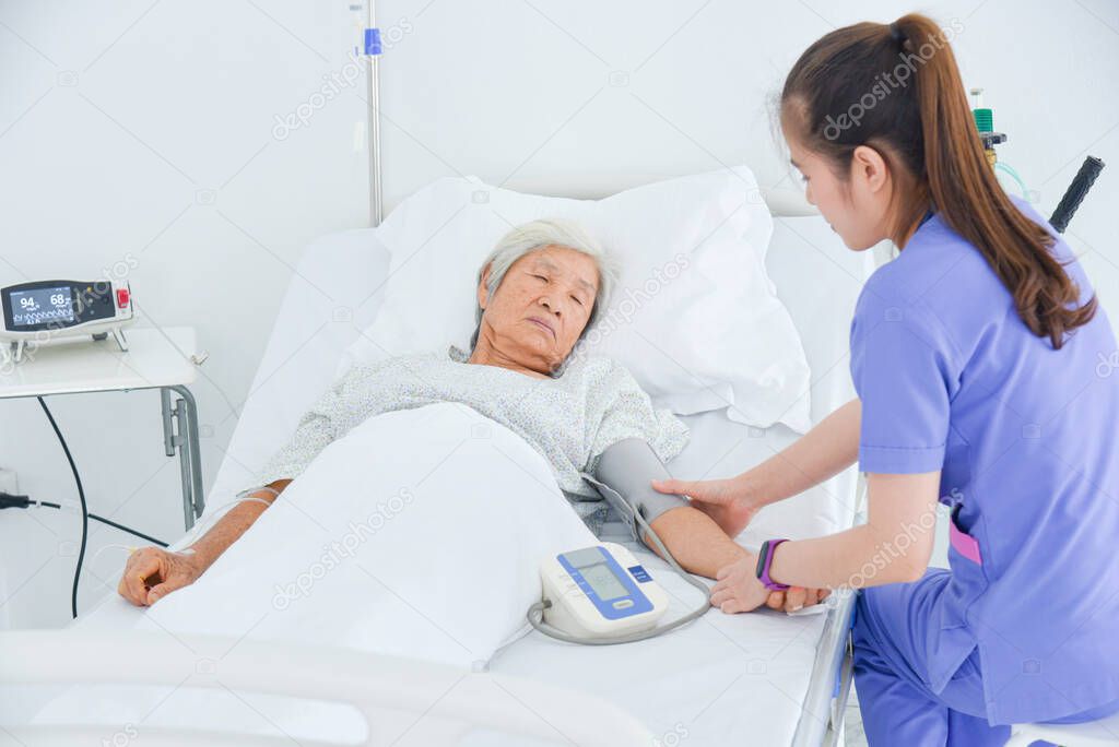 Senior asian female patient lying on bed while nurse measuring blood pressure in hospital ward.