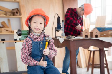 Küçük Asyalı kız miğfer takıyor ve elinde tebessümle çekiç tutuyor marangozhanede oturuyor..