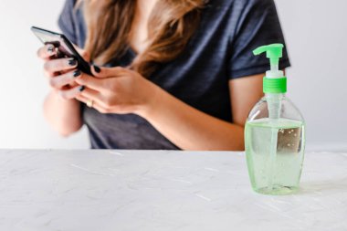 Virüs ve bakterilere karşı iyi hijyen uygulayın. Masanın önünde el dezenfektanlı akıllı telefon kullanan bir kadın.
