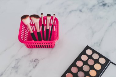 Güzellik endüstrisi ve temel ürünler konsepti, yüz ve göz makyajı için çeşitli makyaj fırçaları içeren pembe alışveriş septi, çıplak tonlu göz falı paleti.