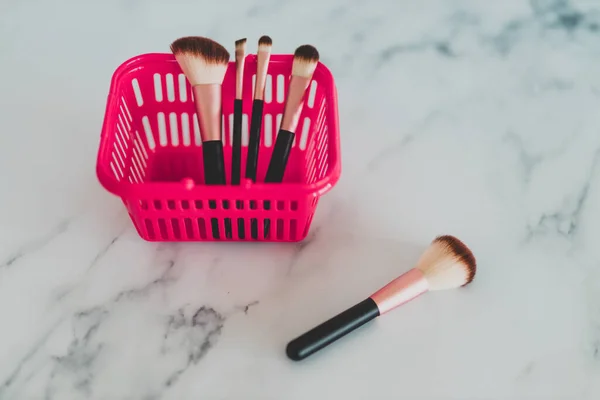beauty industry and essential products concept, pink shopping basket with variety of make-up brushes in it for face and eye make-up and one brush outside in the foreground