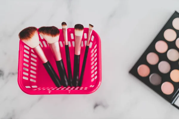 beauty industry and essential products concept, pink shopping basket with variety of make-up brushes in it for face and eye make-up next to eyeshadow palette with nude tones