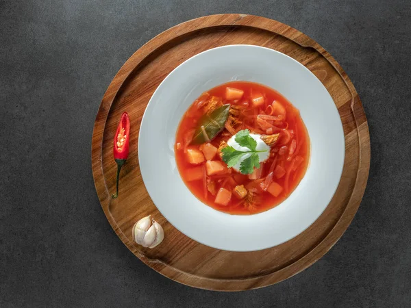 Red ukrainian borscht in white plate on wooden plate with red pepper and garlic on the dark background