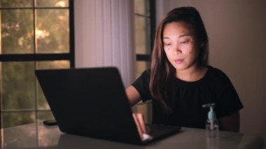 Koronavirüs bulaşmasın diye laptopu ıslak mendil ve mendille temizleyen Asyalı kadın. Sosyal uzaklık ve laptopa odaklanma. El jeli dezenfektanı masaya koy..