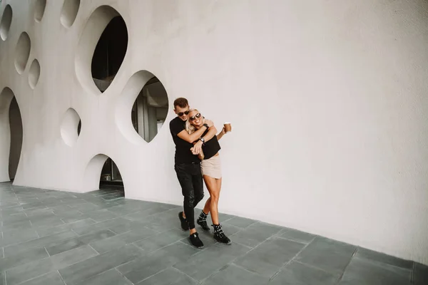 Sweet couple walking along the nice building white wall facade hugging each other. Blonde lady holding coffee in hand with the smiling guy. Street fashion outdoor photography.