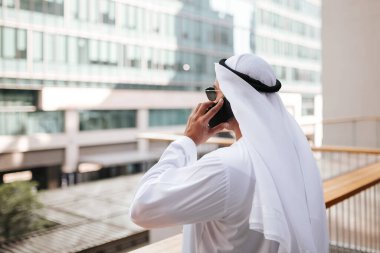 Emirati 'nin şehir emirliklerinde geleneksel kandura giyen adamı akıllı telefonla konuşuyor. Balkondan bakan bina. Arap dergisi için fotoğraf konsepti.