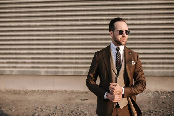 Business man in brown suit and tie wearing sunglass in daytime black wall background. Mans business wear fashion model photoshoot outdoor for magazine cover or clothing images