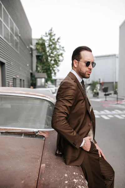 Business man in brown suit and tie wearing sunglass in daytime black wall background. Mans business wear fashion model photoshoot outdoor for magazine cover or clothing images