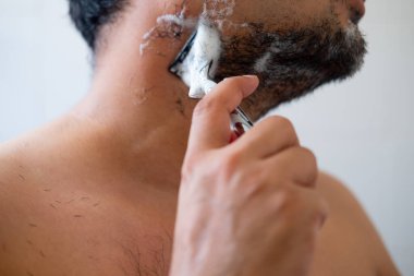 Yanaklarında köpük sabunu olan genç adam duruyor ve bıyığını yüzünü tıraş ederek kesiyor..