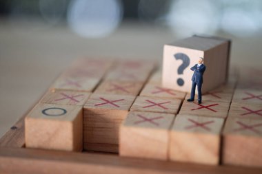 Koyu mavi takım elbiseli minyatür iş adamı tahta blokta soru işaretiyle duruyor ve iş hakkında düşünüyor.