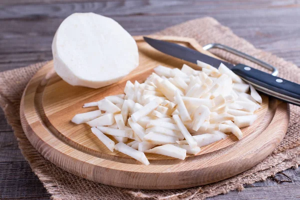 Root celery cut into small pieces with the half of the whole celery root on a wood cutting board on a light wooden background. Step by step cooking. — Stock Photo, Image