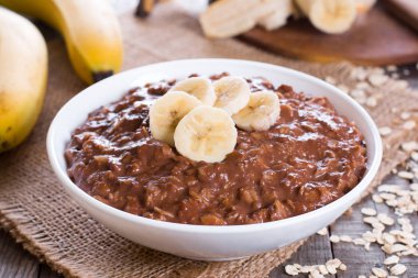 Yatay bir konumda ahşap arka plan üzerinde beyaz bir kase içinde olgun bir muz dilimleri ile kahvaltıda çikolata yulaf ezmesi.