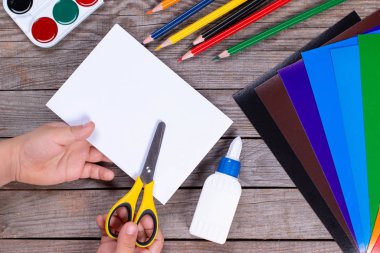 Çocuk bir kağıt şerit kare tutuyor ve onu kesiyor. Çocuk el işi yapıyor. Tahta masaya yapıştırıcı, kağıt, makas. Çocuk sanat projesi, çocuklar için bir zanaat..