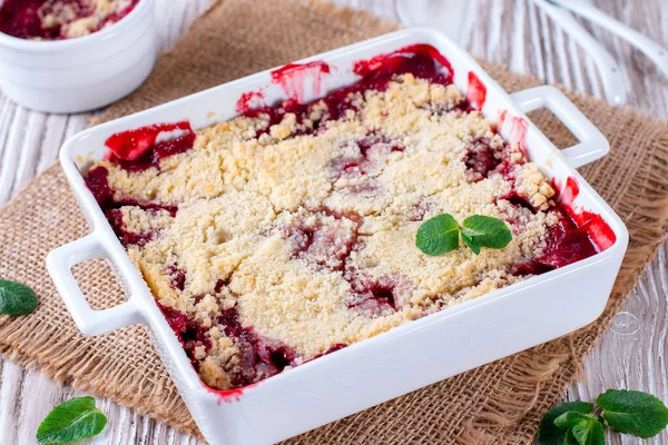 Crumble with strawberries in white dish on white wooden background