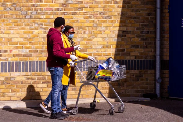 Homerton, Londra, İngiltere, Nisan 7, 2020: Güneşli bir günde dışarıda bir dükkanın önünde kuyrukta bekleyen iki kişi ve Corona zamanlarında maske ve eldiven takan iki kişi