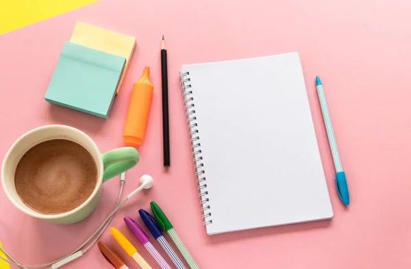 Sketch pad (notebook), multi colored pens, mug with coffee and headphones on the pink background. Stationery in top view design with text space.