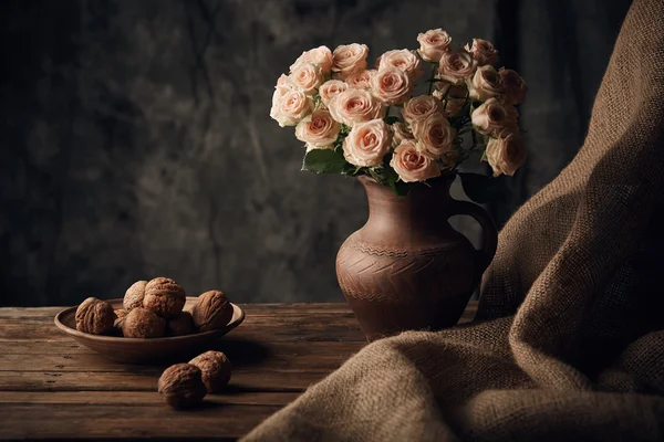 Beautiful still life with light roses and walnuts on wooden table with brown sacking — Stock Photo, Image