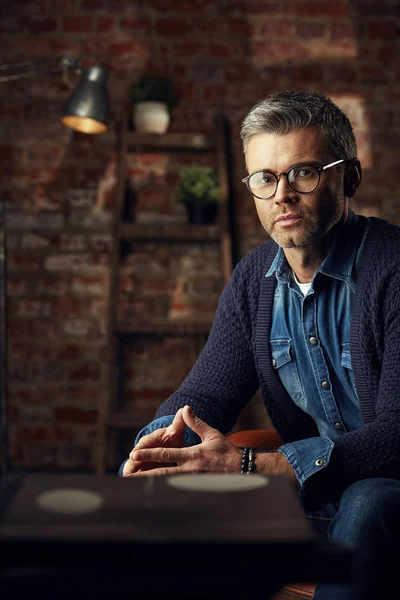 Pria modern dengan rambut abu-abu dan kacamata duduk di loteng studio di sofa dan melihat kamera — Stok Foto