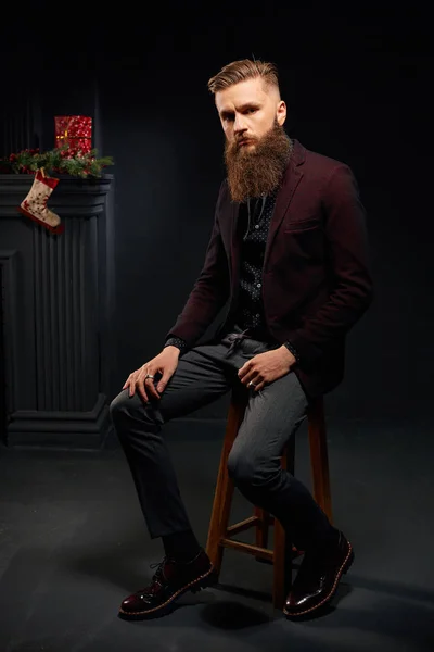 Красивий стильний чоловік з бородою сидить у темній студії з різдвяною шкарпеткою на фоні — стокове фото