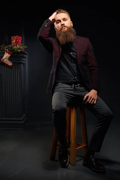 Pemuda model laki-laki dengan jenggot duduk di studio gelap dengan kaus kaki Natal di latar belakang — Stok Foto