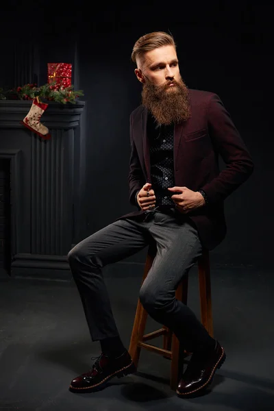 Handsome young man with beard sitting in dark studio with christmas sock on background — Stock Photo, Image