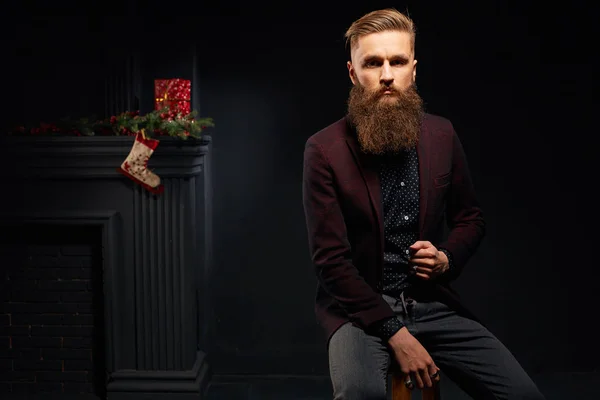 Modelo macho con barba sentado en estudio oscuro con calcetín de Navidad en el fondo —  Fotos de Stock