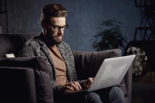 Retrato de un hombre de negocios elegante y guapo vestido de gris y gafas, con barba, sentado en muebles de lujo en habitación oscura — Foto de Stock