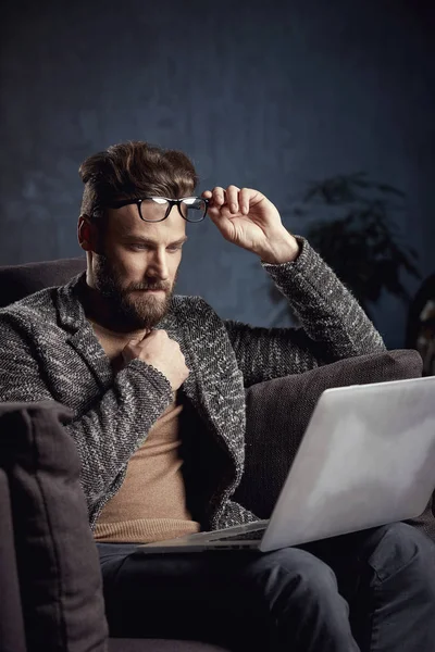 Retrato de un hombre de negocios elegante y guapo vestido de gris y gafas, con barba, sentado en muebles de lujo con portátil — Foto de Stock