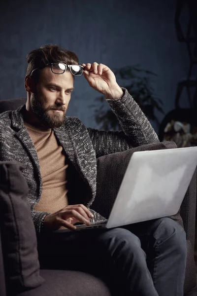 Retrato de un hombre de negocios elegante y guapo vestido de gris y gafas, con barba, sentado en muebles de lujo con portátil — Foto de Stock