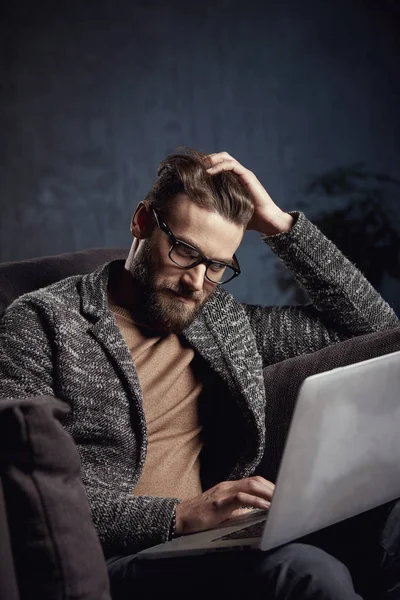 Atractivo serio elegante hombre de negocios moderno con gris y gafas, con barba, sentado en muebles caros con ordenador portátil — Foto de Stock