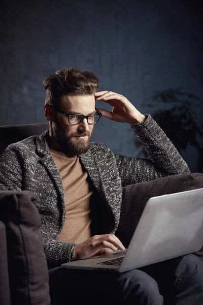 Atractivo serio elegante hombre de negocios moderno con gris y gafas, con barba, sentado en muebles de lujo con ordenador portátil — Foto de Stock