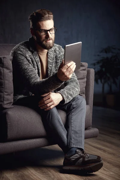 Atractivo serio elegante hombre de moda con gris y gafas, con barba, sentado y leyendo en un elegante sofá en el interior oscuro de moda — Foto de Stock