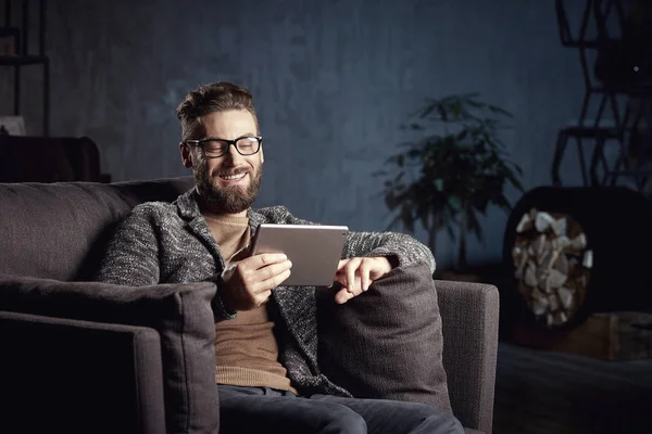 Hombre clásico elegante sonriente lindo que usa gris y gafas, con barba, sentado y leyendo en muebles elegantes en el interior oscuro de moda — Foto de Stock