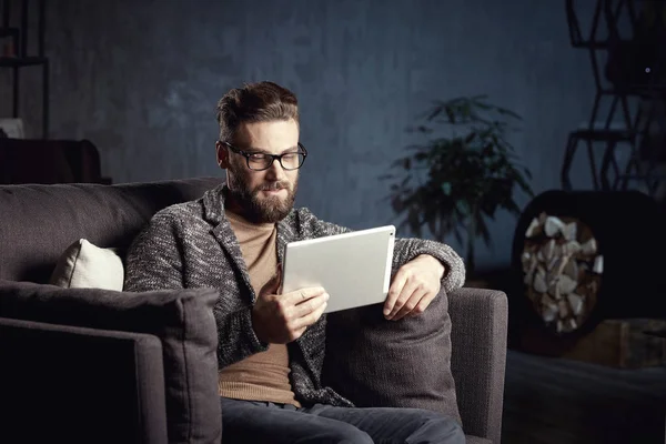 Hombre clásico elegante serio guapo que usa gris moderno y gafas, con barba, sentado en muebles elegantes en el interior oscuro de moda con el ordenador portátil — Foto de Stock