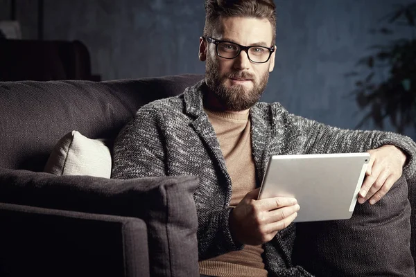 Hombre clásico elegante serio guapo que usa gris moderno y gafas, con barba, sentado en muebles elegantes en el interior oscuro de moda con el ordenador portátil — Foto de Stock