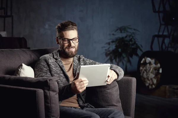 Handsome attractive elegant classic man wearing trendy grey and glasses, with beard, sitting on stylish furniture in fashionable dark interior with laptop