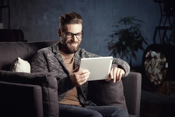 Handsome attractive elegant classic man wearing trendy grey and glasses, with beard, sitting on stylish furniture in fashionable dark interior with laptop