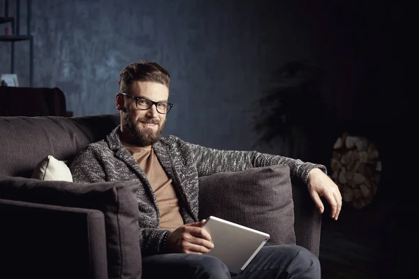 Hombre clásico elegante atractivo guapo que usa gris moderno y gafas, con barba, sentado en muebles elegantes en el interior oscuro de moda con el ordenador portátil — Foto de Stock