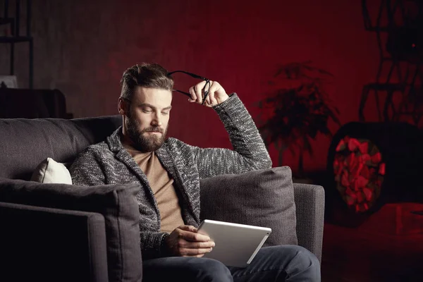 Красивый привлекательный элегантный классический мужчина в модных сером и очках, с бородой, сидя на стильной мебели в модном темно-красном интерьере с ноутбуком — стоковое фото